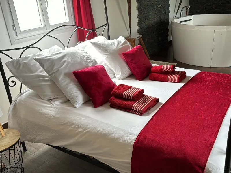 Chambre cosy avec lit en fer forgé, linge blanc rehaussé de rouge et baignoire balnéo ronde