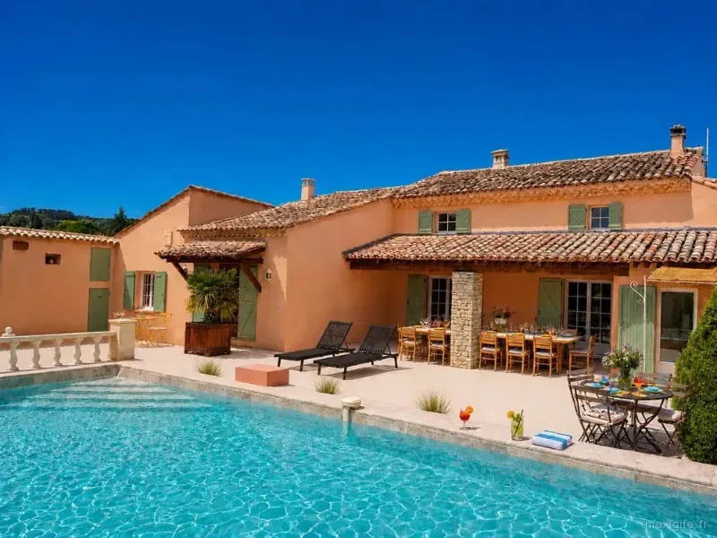 Façade ocre du mas aux volets verts avec terrasse, table dressée et piscine turquoise au soleil