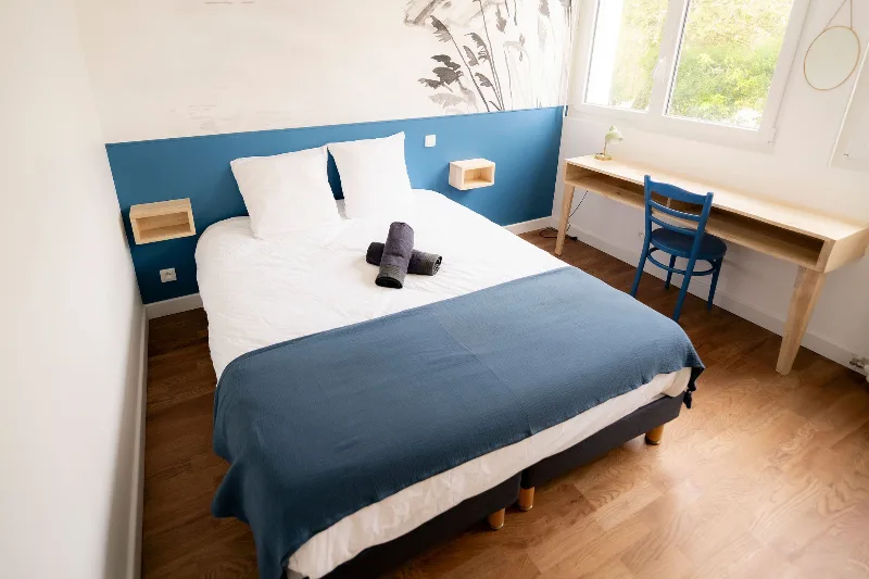 Chambre au mur bleu avec lit double, bureau en bois clair, chaise et fenêtre sur la nature
