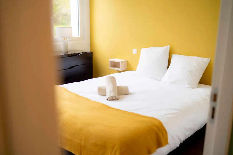 Chambre lumineuse au mur jaune avec lit double, commode foncée et fenêtre donnant sur la verdure
