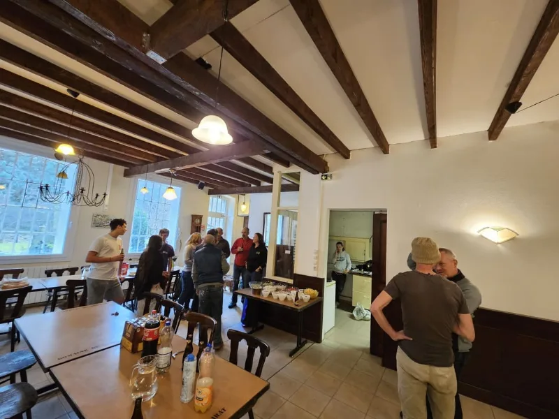 Grande salle a manger sous poutres apparentes avec tables, convives et cuisine attenante