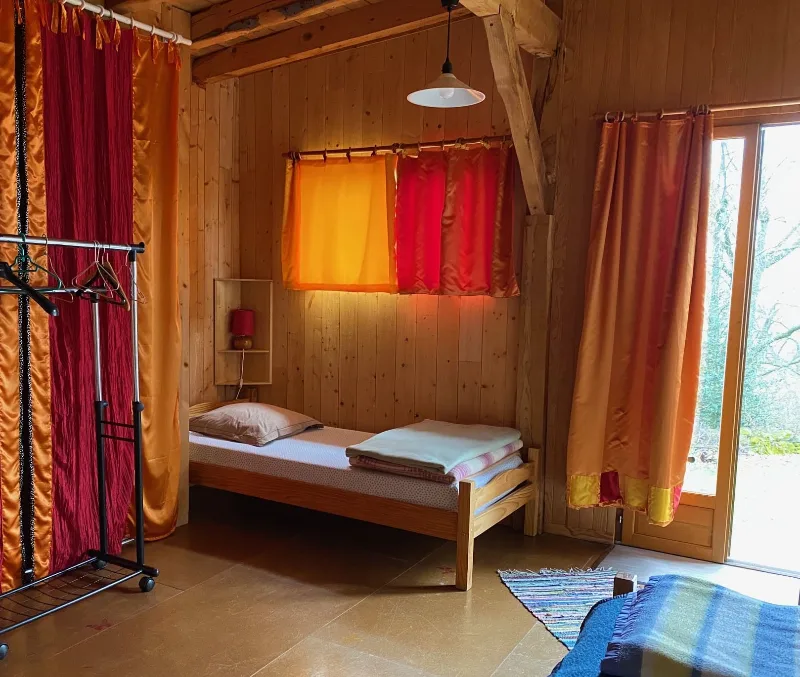 Chambre chaleureuse en lambris de bois avec lit simple, rideaux oranges et baie vitree sur jardin