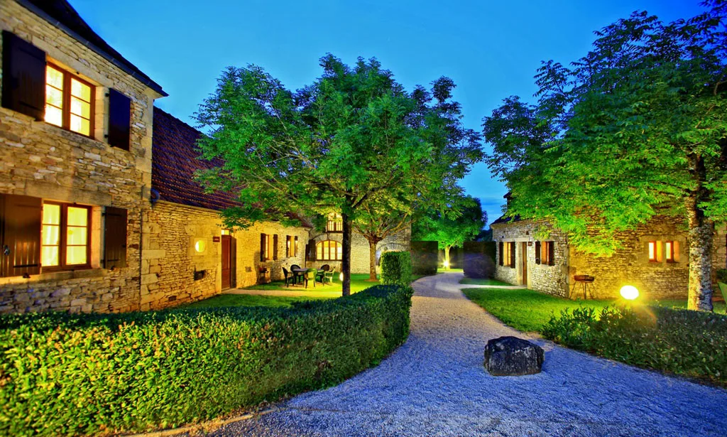 Gîte de groupe Combas Village de Gîtes à Saint-Crépin-Carlucet — Gîtes en pierre illuminés à la tombée de la nuit avec allée gravillonnée et arbres éclairés, capacité 70 personnes