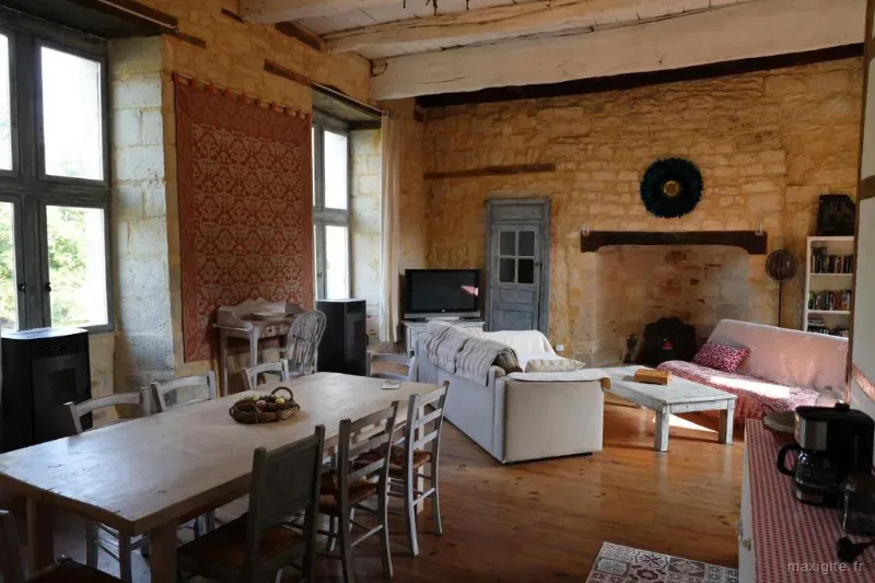 Grand séjour en pierre avec longue table en bois, cheminée ancienne, canapé et belles fenêtres