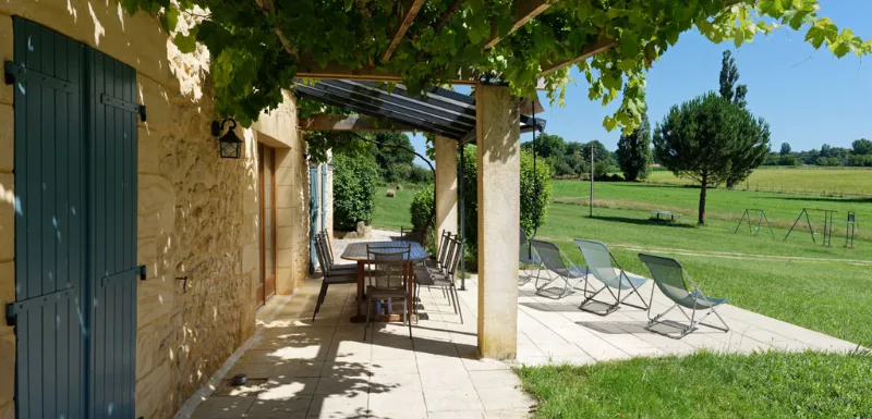 Terrasse ombragée sous pergola de vigne le long de la façade en pierre, ouverte sur la campagne