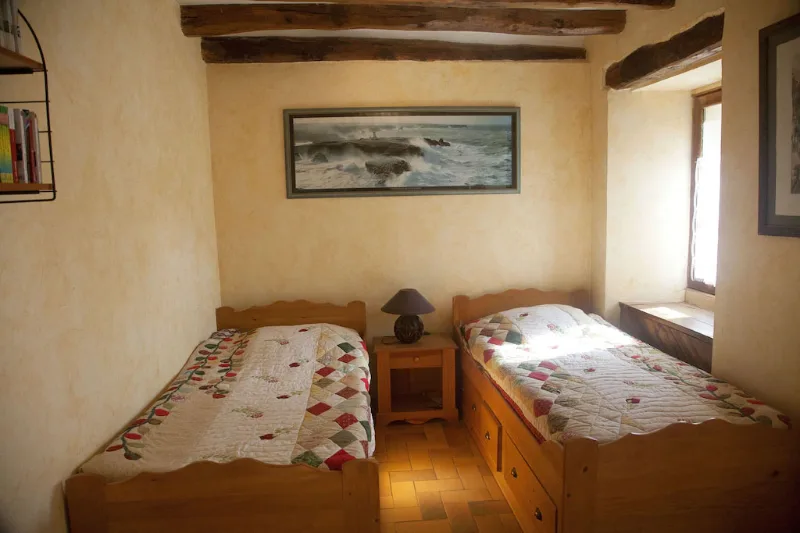 Chambre sous poutres anciennes avec deux lits simples, sol en tomettes et fenêtre lumineuse