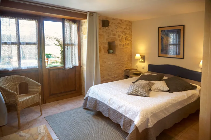 Chambre chaleureuse avec lit double, mur en pierre apparente et porte-fenêtre ouverte sur la nature