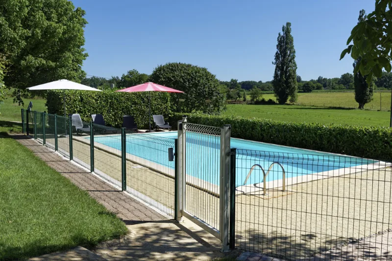 Piscine clôturée avec parasols colorés et pelouse verdoyante bordée d'une haie en arrière-plan