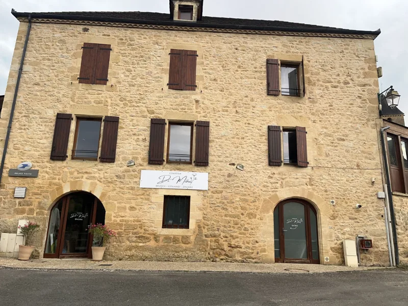 Façade en pierre de la bâtisse à trois niveaux avec volets bois et entrées en arc sur la place