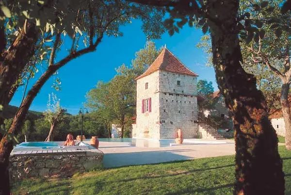 Pigeonnier en pierre et bâtisses anciennes dominant la piscine, jardin verdoyant et ciel bleu