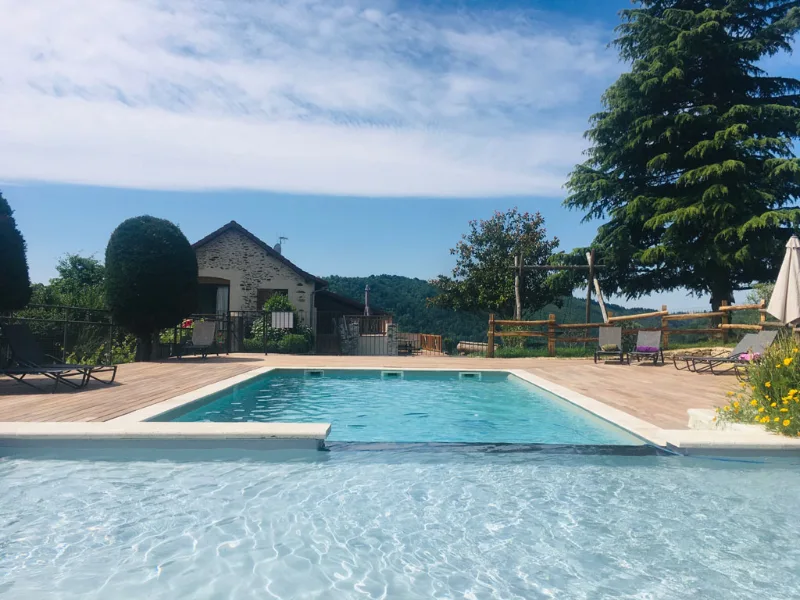 Piscine extérieure et terrasse en bois avec transats devant la maison en pierre et collines boisées