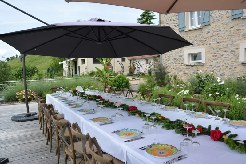 Longue table dressée pour un repas de groupe sur la terrasse, parasols et façade en pierre