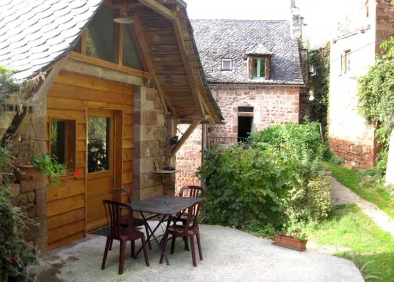Façade en pierre rouge avec extension bois et terrasse meublée d'une table et chaises de jardin