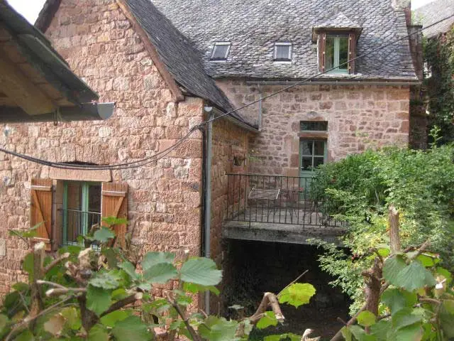 Bâtisse traditionnelle en pierre rouge avec volets bois, balcon en fer forgé et toiture en lauzes