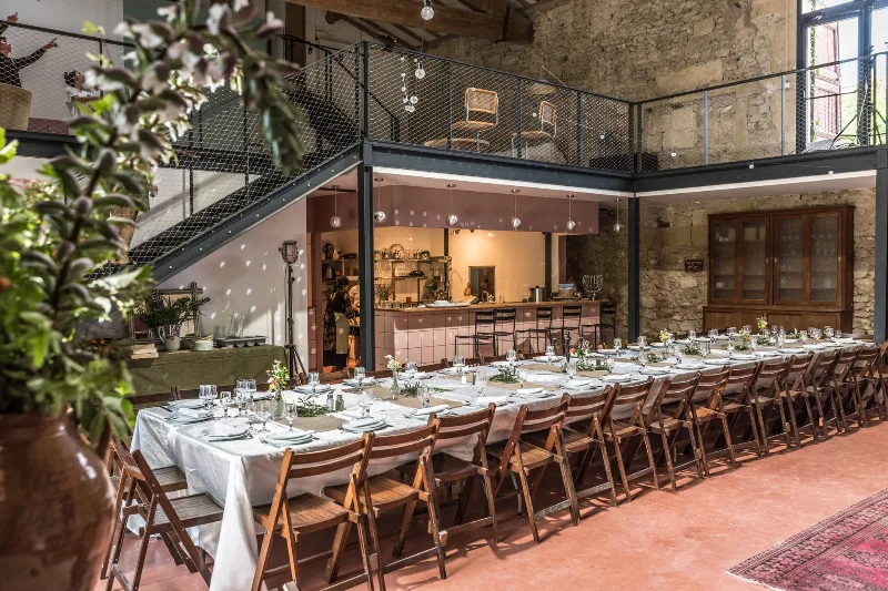 Grande salle voûtée en pierre avec longue tablée dressée, mezzanine et cuisine ouverte au fond