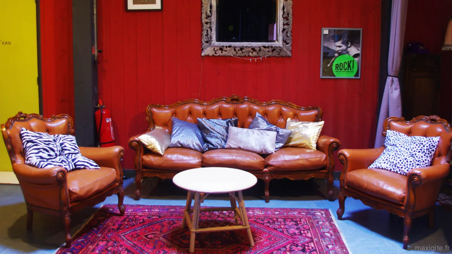 Gîte de groupe Gîtes de l'École à Sizun Saint Cadou — Salon vintage avec canapé en cuir caramel, coussins dépareillés et mur rouge, capacité 22 personnes