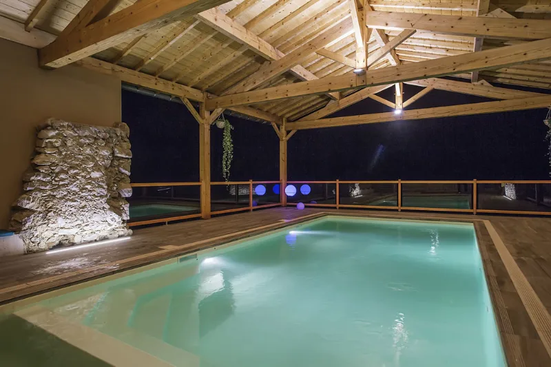 Piscine couverte illuminée de nuit sous charpente bois et mur en pierre
