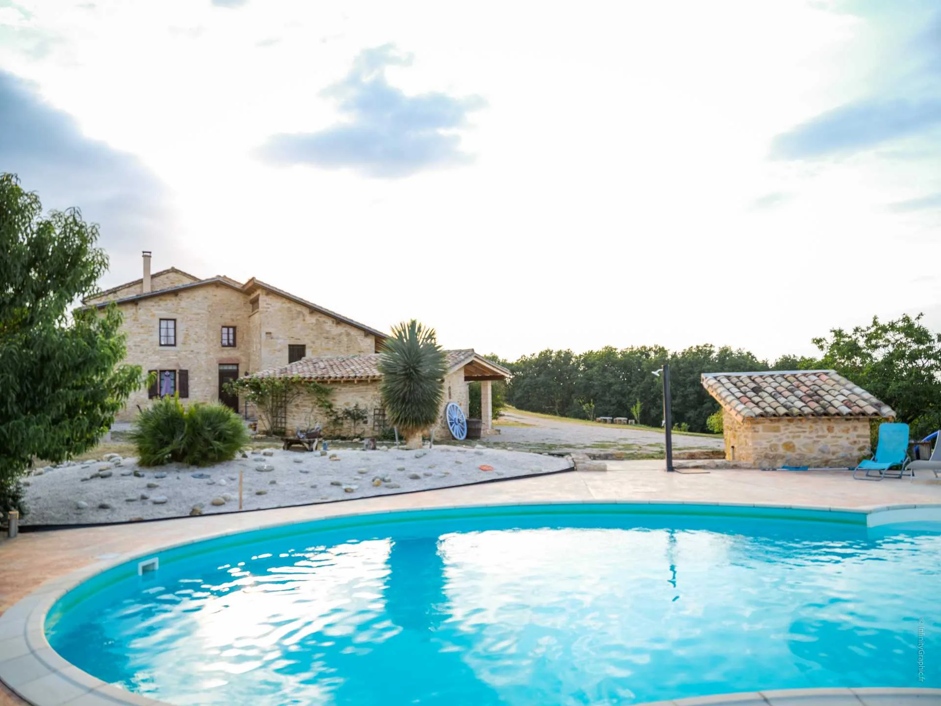 Gîte de groupe Domaine du Colombier à Larroque — Piscine ronde devant corps de ferme en pierre sous grand ciel nuageux, capacité 29 personnes