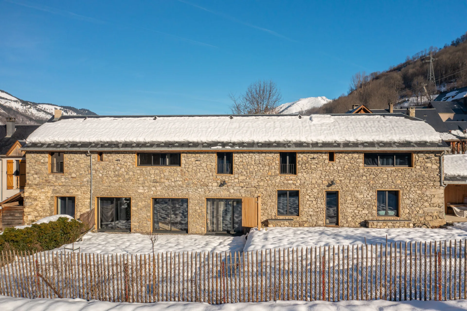 Gîte de groupe Chalet de Soos à Loudenvielle — Façade en pierre du chalet sous ciel bleu avec vue sur les sommets pyrénéens enneigés, capacité 12 personnes