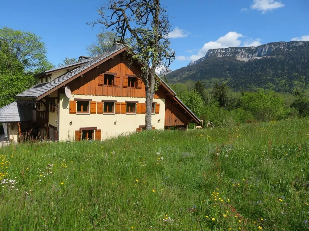Gîte de groupe Domaine de la Sapinière à Fillière — Grand chalet en bois et crépi clair au milieu d'une prairie fleurie, face aux falaises calcaires de Haute-Savoie, capacité 32 personnes