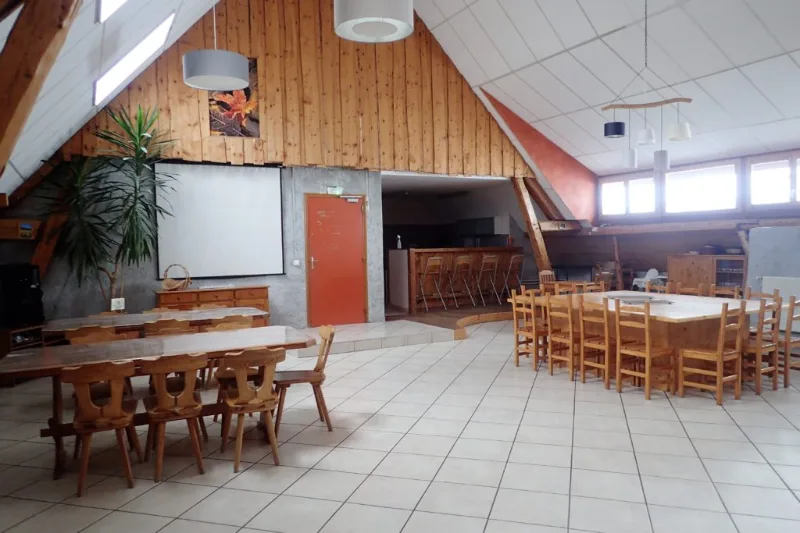 Grande salle commune avec tables en bois, chaises en sapin et écran de projection