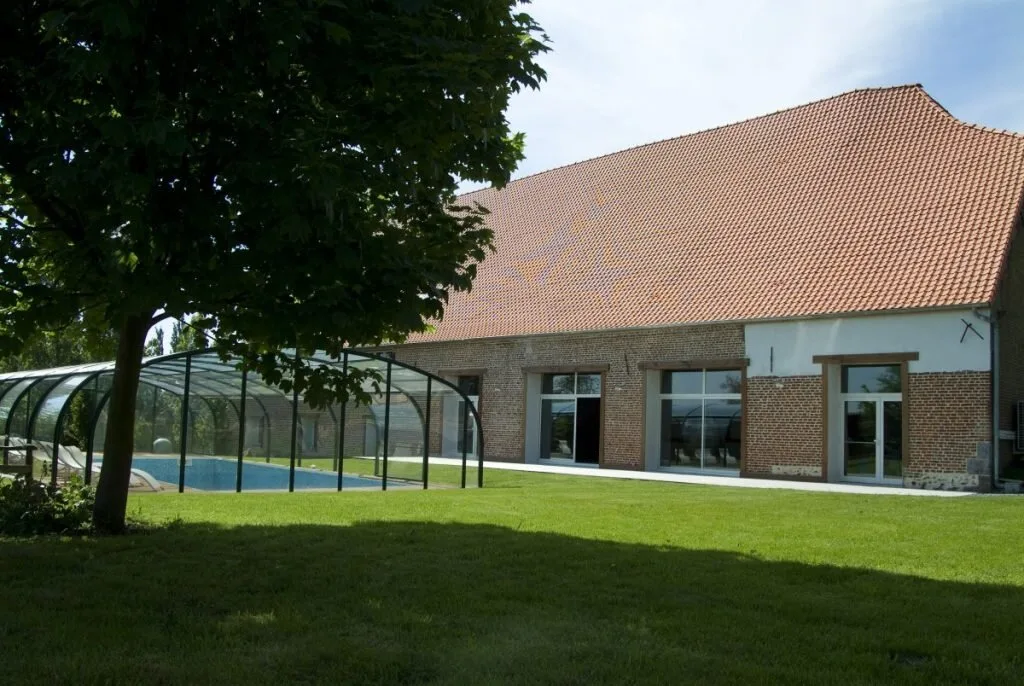 Gîte de groupe La Ferme de Saint-Humbert à Salesches — Façade en briques et toiture rouge de la ferme avec piscine couverte et jardin arboré, capacité 30 personnes