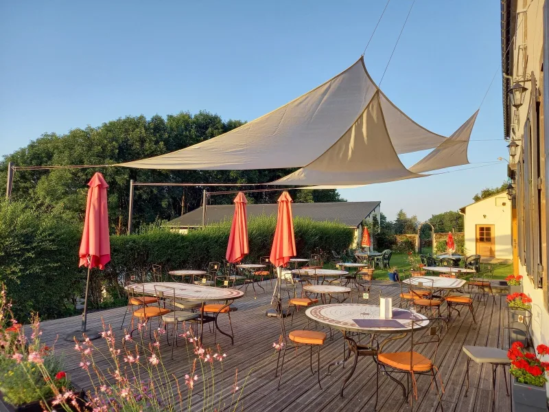 Terrasse extérieure avec tables et chaises, ombragée par des arbres devant la façade en pierre