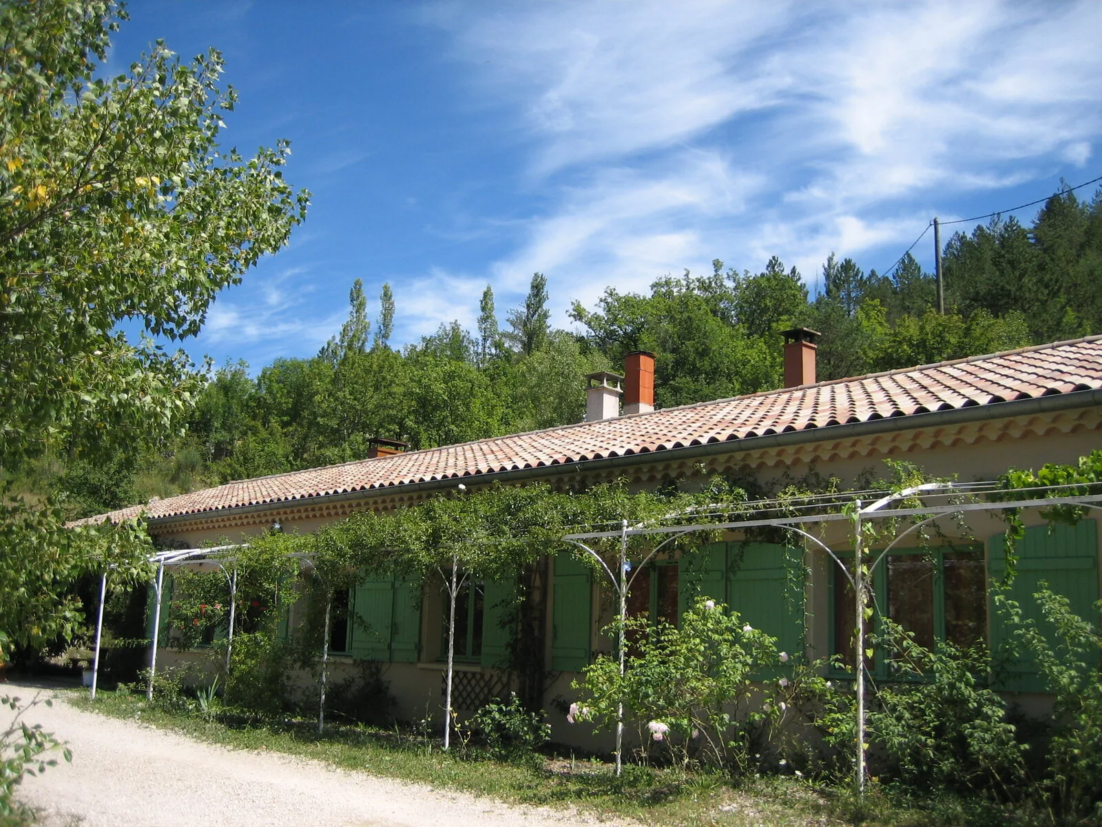 Gîte de groupe Le Présent Simple à Cornillon-sur-l'Oule — Façade de la maison commune aux volets verts et toiture provençale, entourée de pergolas fleuries, capacité 23 personnes