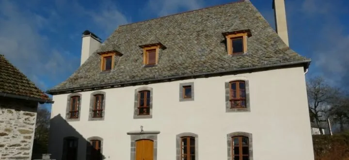 Gîte de groupe Gites de la Forêt à Arpajon-sur-Cère — Vue de face de la bâtisse en pierre du Cantal sous un ciel bleu, deux niveaux, capacité 18 personnes