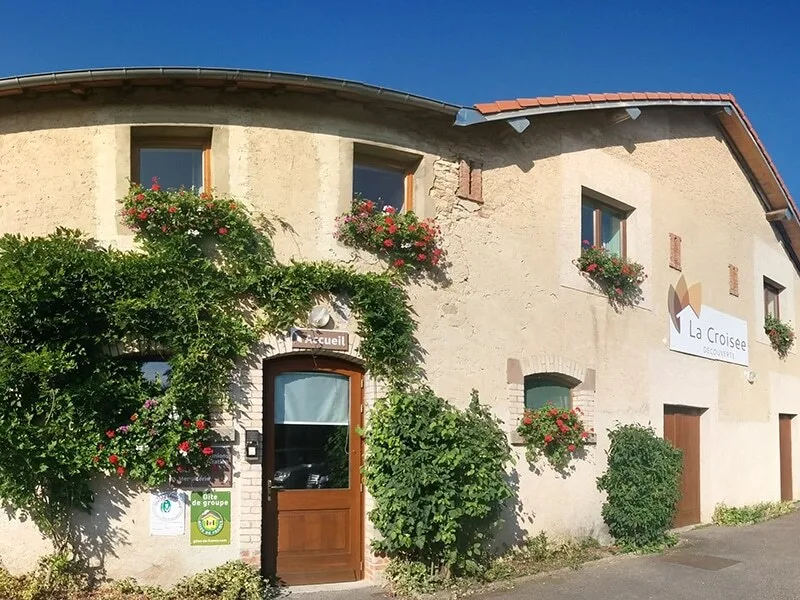 Façade en pierre de l'ancienne ferme avec géraniums aux fenêtres et enseigne du gîte
