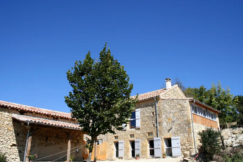 Façade en pierre gasconne avec volets bleus, toits de tuiles et grand arbre