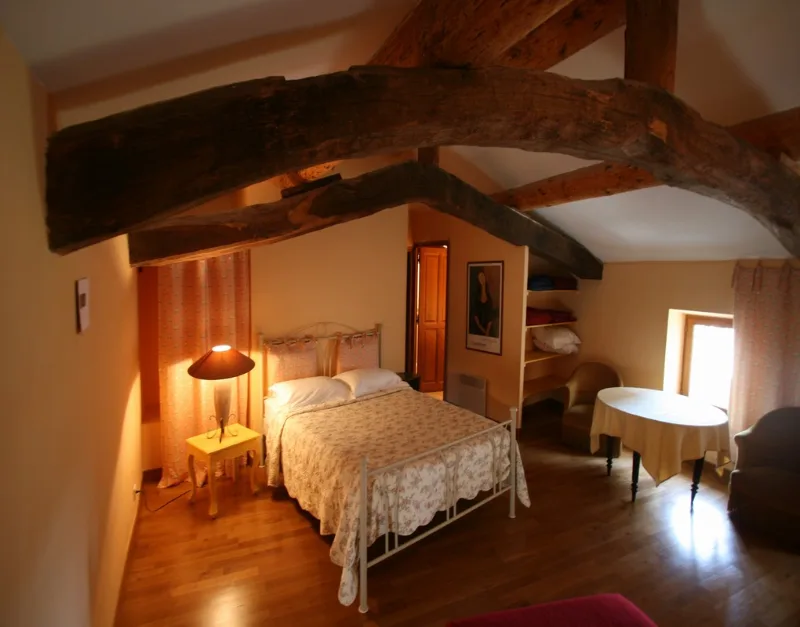 Chambre mansardée avec poutres apparentes, lit en fer forgé et petit salon de lecture