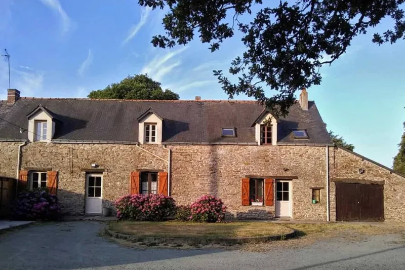 Longère bretonne en pierre avec volets rouges, lucarnes et grand parking devant la maison
