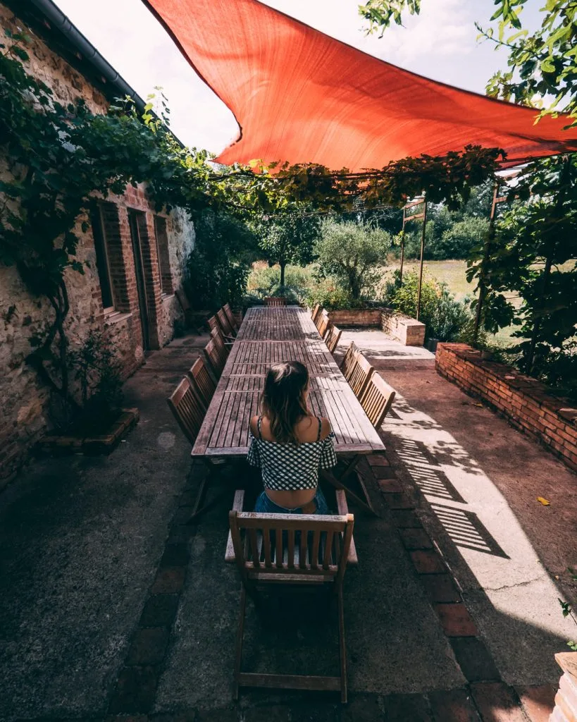 Gîte de groupe Gite Anthuai à Mérigny — Longue table en bois sous une pergola et un voile ombragé, contre la ferme en pierre, capacité 25 personnes