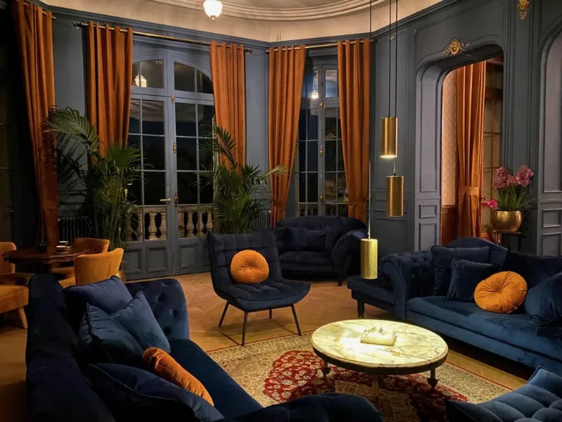Salon bleu nuit avec fauteuils veloutes, coussins orange et portes-fenetres sur le parc