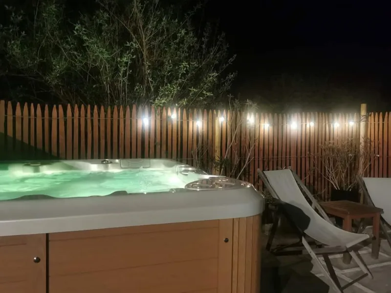 Jacuzzi exterieur eclaire en soiree, entoure d'une cloture en bois