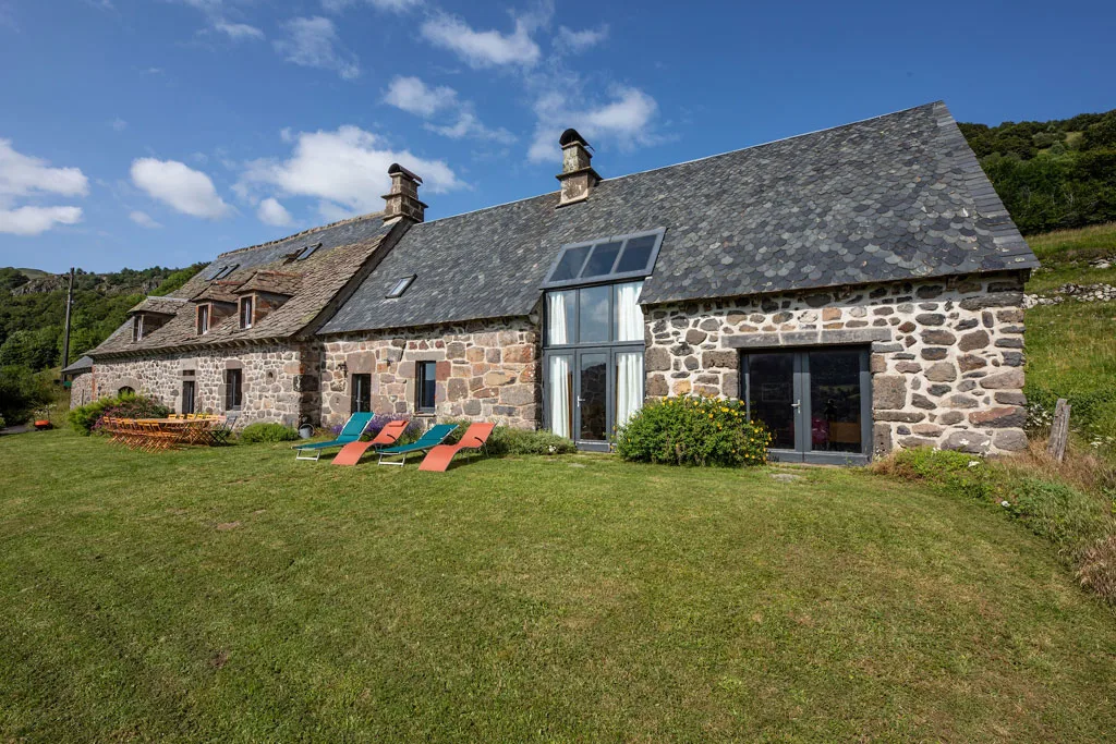 Gîte de groupe L'Impradine à Le Falgoux — Façade en pierre du gîte avec toit d'ardoise, transats colorés et jardin verdoyant, capacité 15 personnes