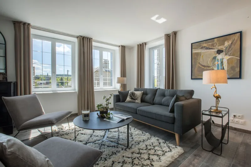 Salon lumineux avec canapés gris, tapis berbère et grandes fenêtres sur le village