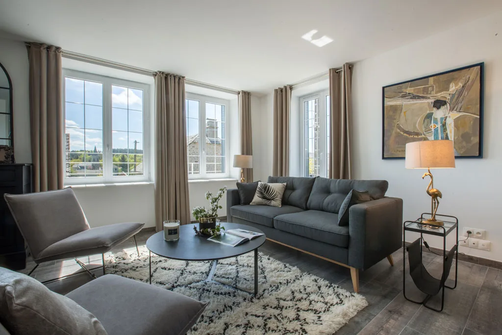 Salon lumineux avec canapés gris, tapis berbère et grandes fenêtres sur le village