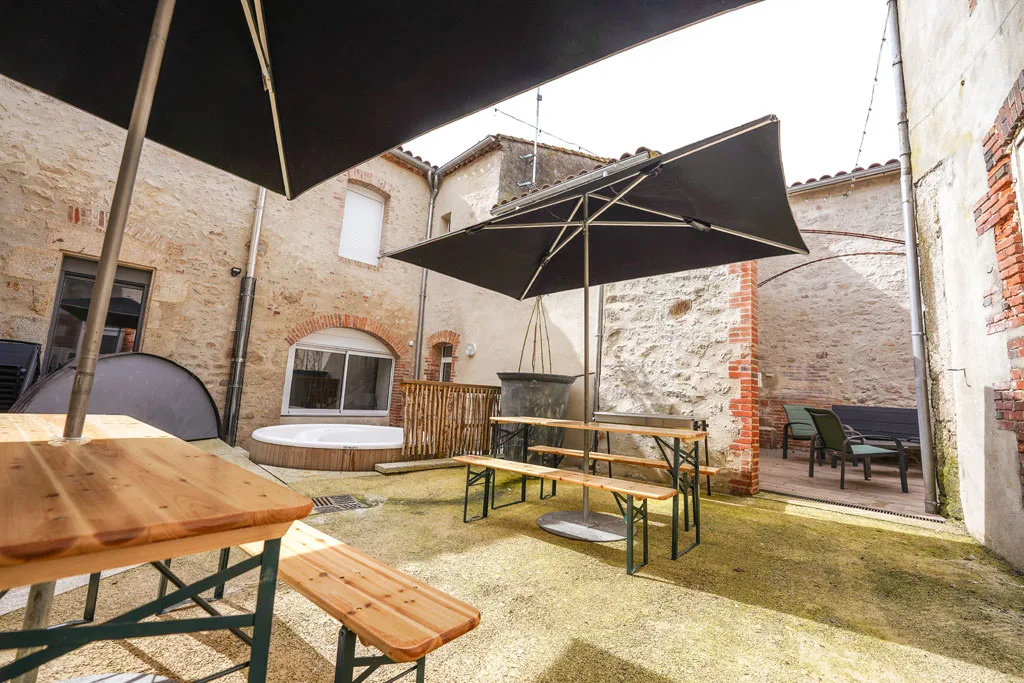 Gîte de groupe Le Patio à Saint-Amand-sur-Sèvre — Patio extérieur avec bain nordique, tables en bois et parasols pour les repas en plein air, capacité 23 personnes