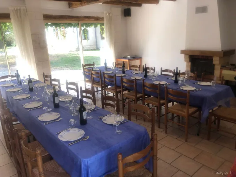 Grande salle à manger avec tables en U dressées, nappes bleues, cheminée et poutres