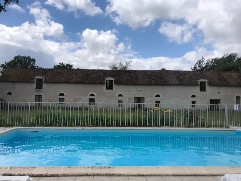 Piscine rectangulaire sécurisée devant longue grange en pierre, ciel d'été nuageux