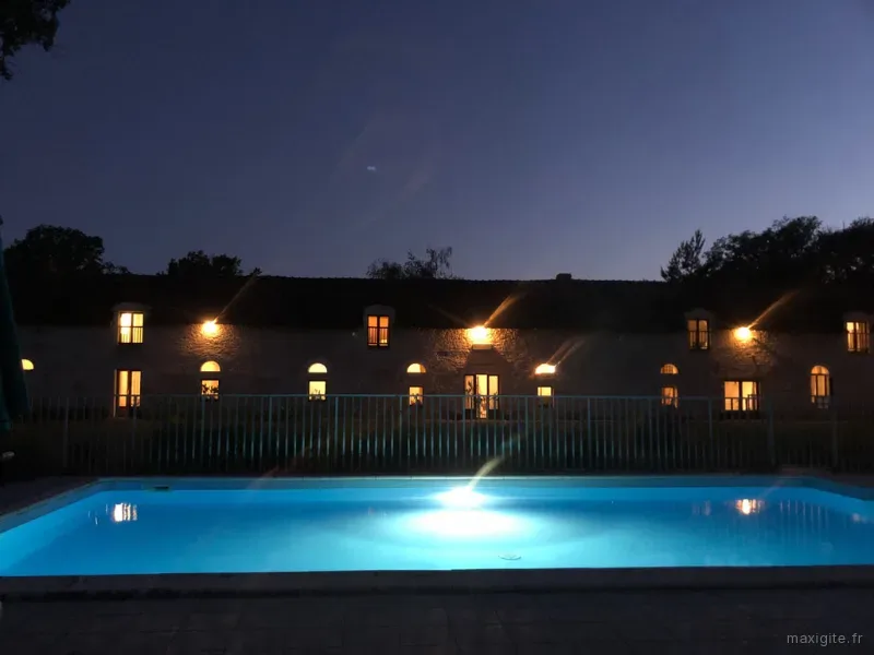 Grange en pierre illuminée de nuit avec piscine éclairée bleue et reflets dans l'eau