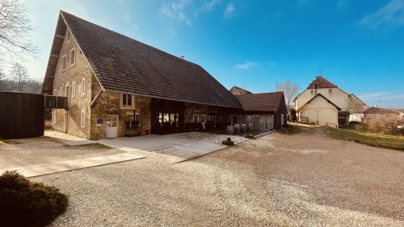 Façade en pierre de la grange rénovée du Moulin Marion, cour gravillonnée et hameau du Sundgau en arrière-plan
