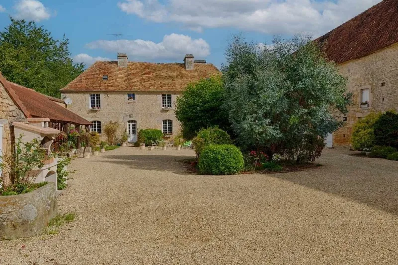 Façade en pierre de la ferme normande avec toiture en tuiles et cour gravillonnée arborée