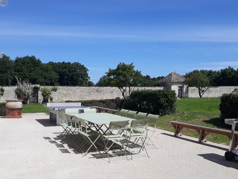 Terrasse extérieure avec grande tablée, chaises et table de ping-pong devant le mur en pierre du domaine