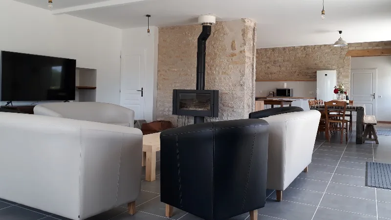 Grand séjour lumineux avec poêle à bois central, mur en pierre apparente et canapés en cuir clair