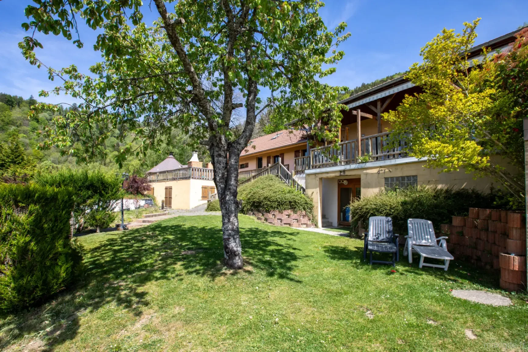 Façade du gîte en pierre vue depuis le jardin, terrasse boisée et chaises longues