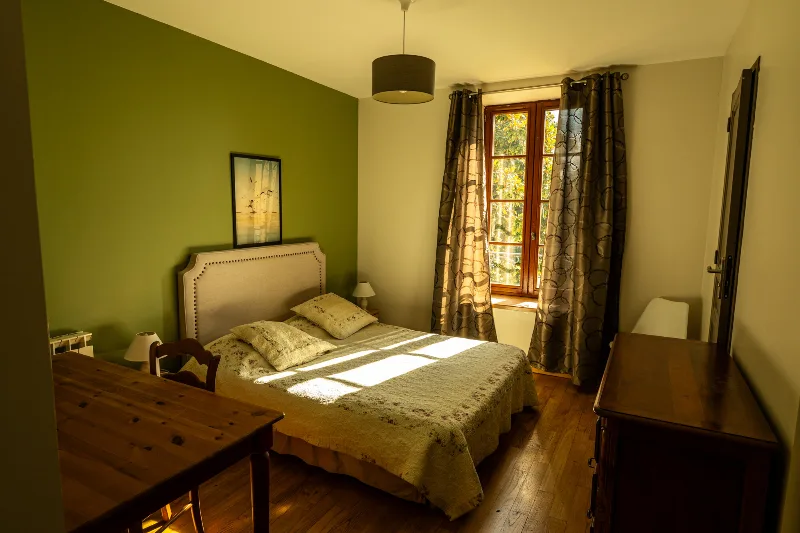Chambre double avec tête de lit capitonnée, mur vert et fenêtre donnant sur les arbres