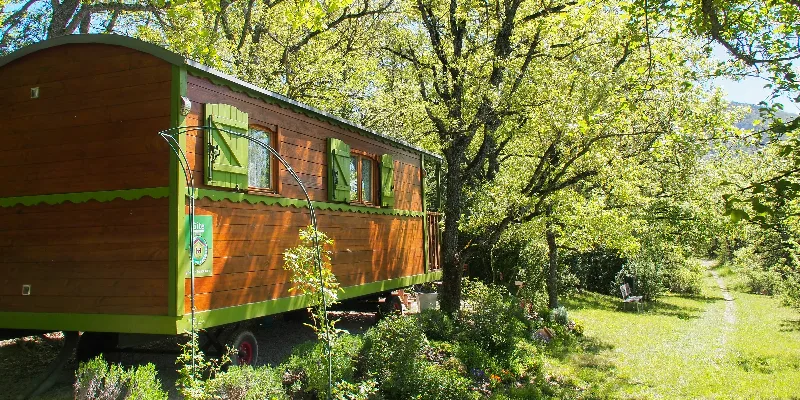 Roulotte en bois avec volets verts nichée sous les arbres, terrasse et jardin fleuri
